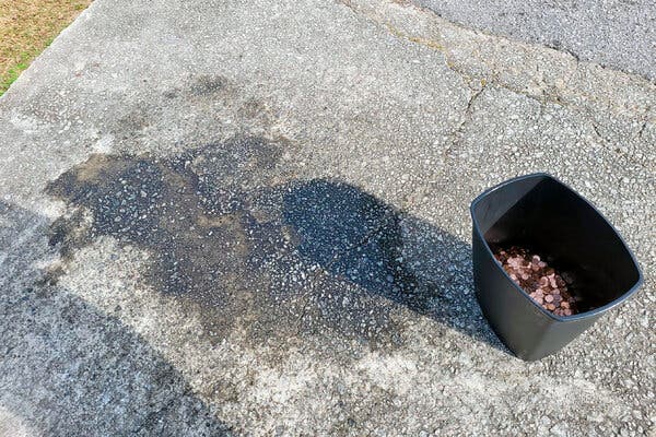 A black plastic can holds a mound of pennies next to the stain on the driveway where they had been left.
